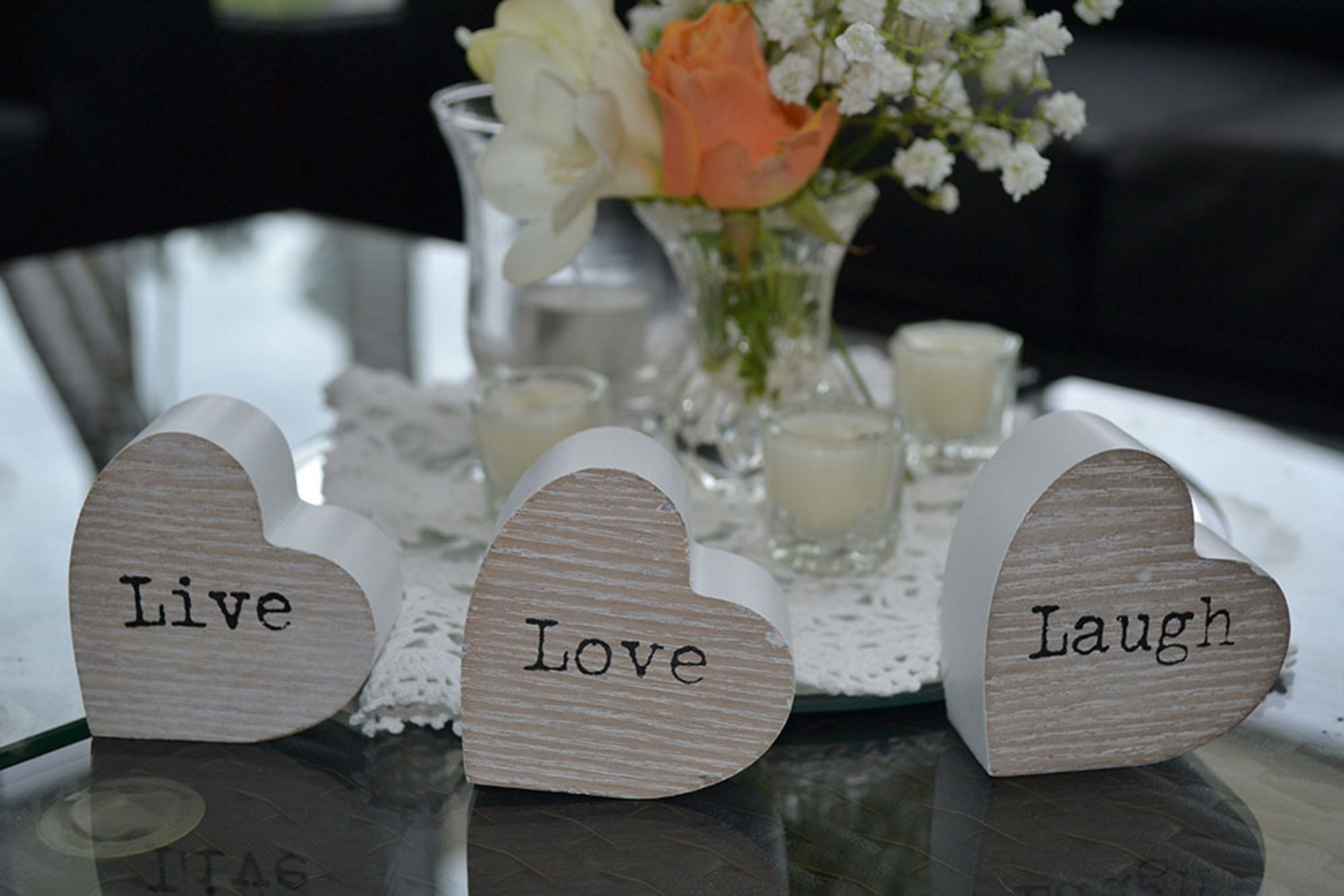 Three wooden heart-shaped signs with words "Live," "Love," and "Laugh" on a table with a lace doily, candles, and a vase with flowers.