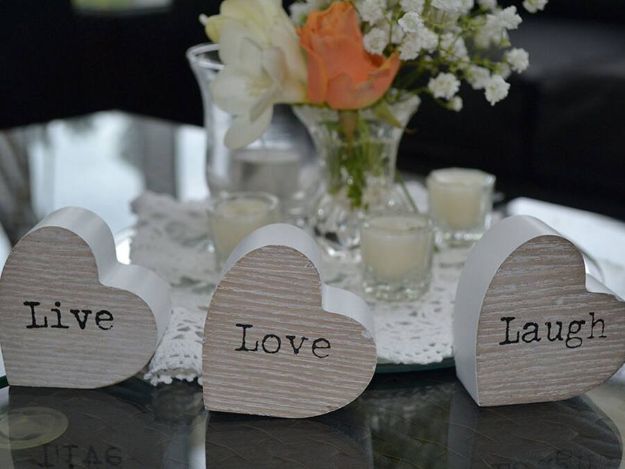 Three wooden heart-shaped signs with words "Live," "Love," and "Laugh" on a table with a lace doily, candles, and a vase with flowers.