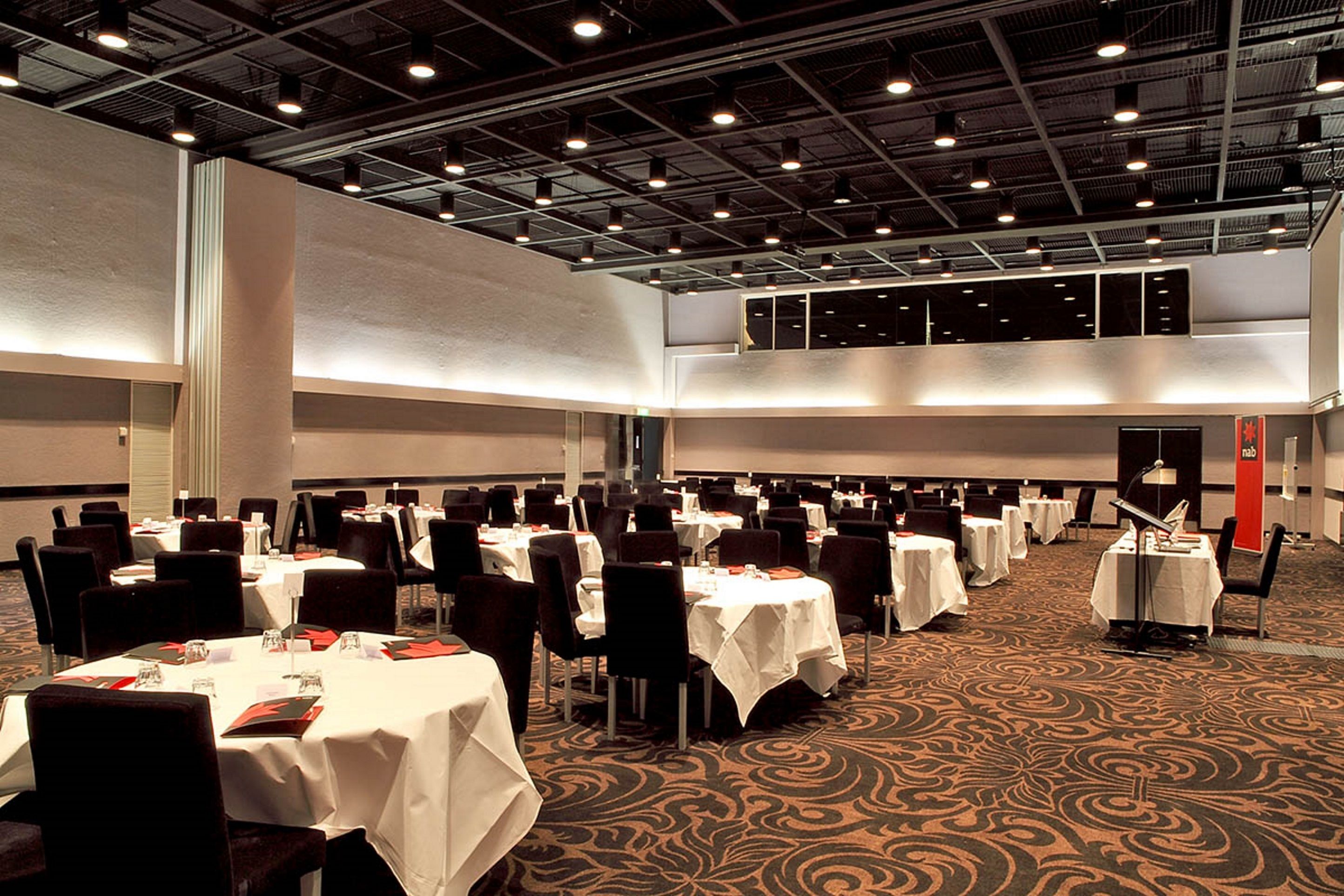 Une grande salle de banquet dotée de nombreuses tables rondes et de chaises, de hauts plafonds avec des poutres apparentes et d’un sol recouvert de moquette.