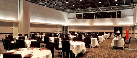 Une grande salle de banquet dotée de nombreuses tables rondes et de chaises, de hauts plafonds avec des poutres apparentes et d’un sol recouvert de moquette.