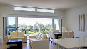 A modern living room with a sofa, a coffee table, and a dining area with chairs and a table. Large windows offer a view of the outdoors. - Opal Cove Resort (Korora)