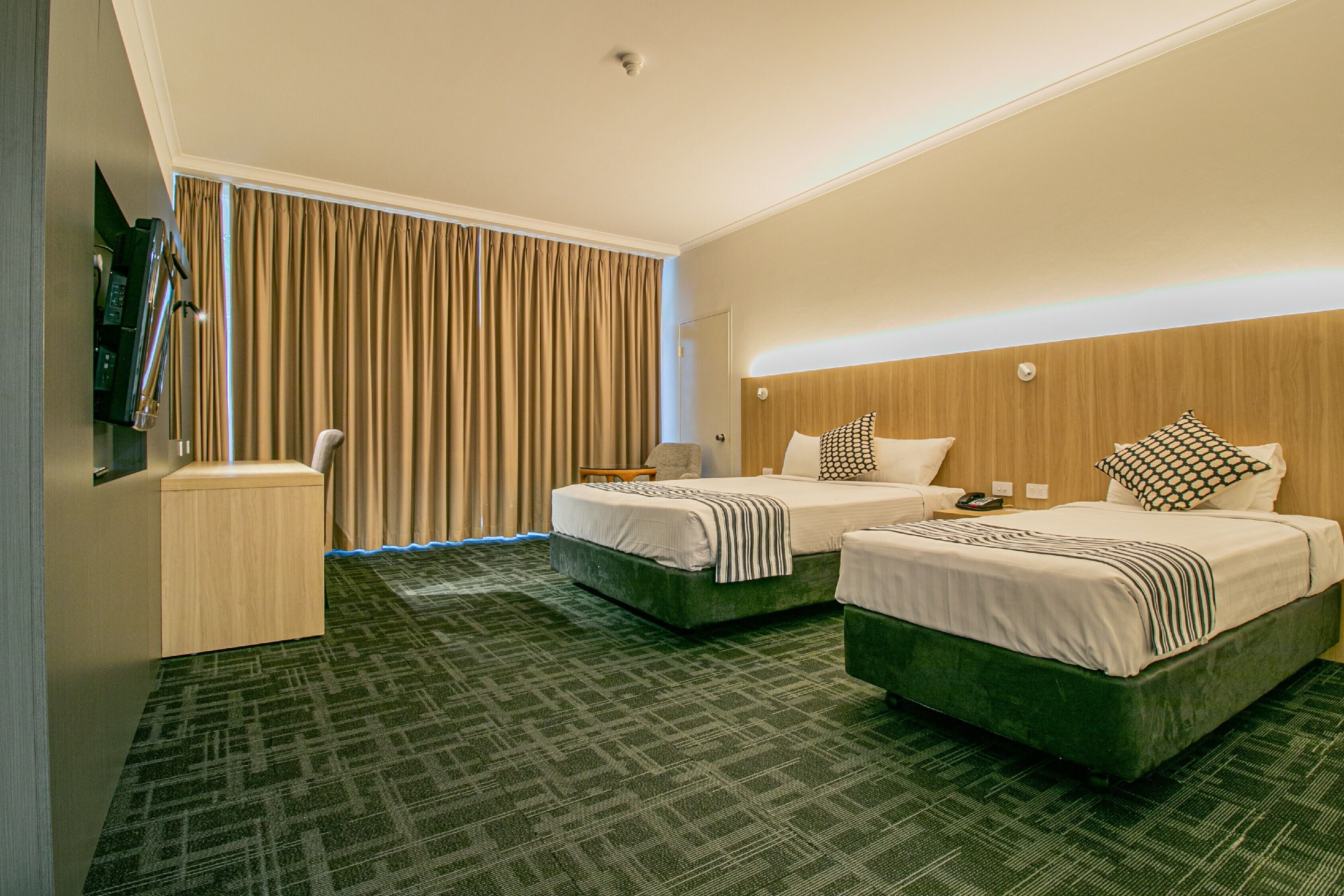 Quarto de hotel com duas camas, uma escrivaninha, uma cadeira, uma televisão e uma janela com cortinas.