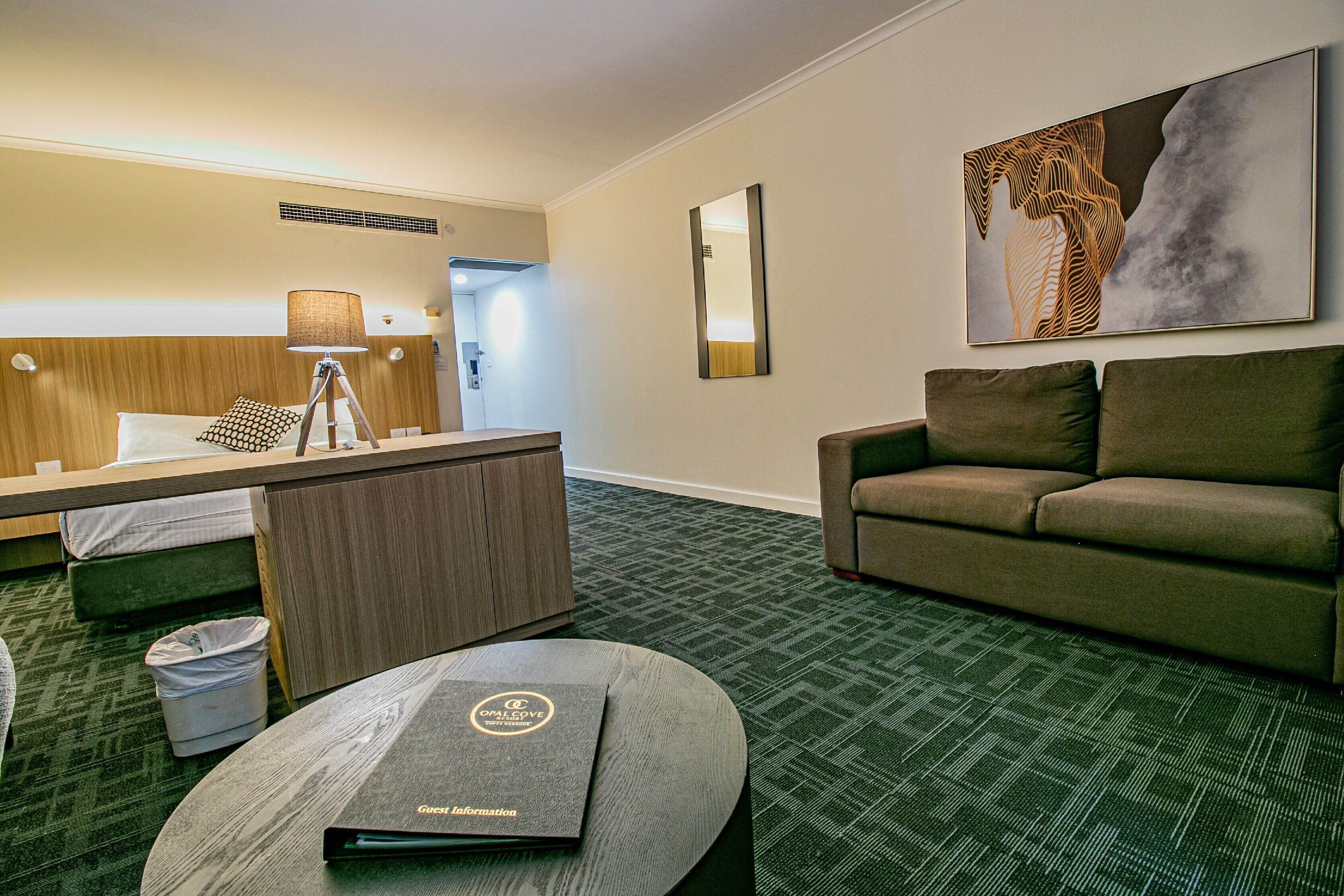 Une chambre d’hôtel avec un lit, un bureau, une lampe, un canapé, une poubelle et un sol recouvert de moquette.