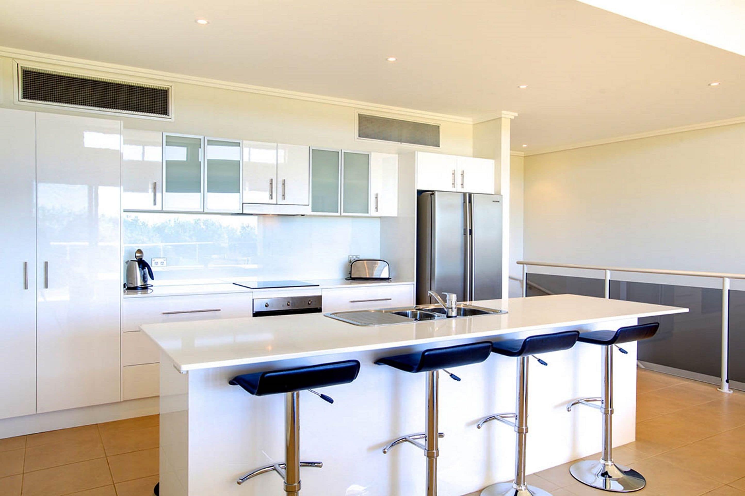 A modern kitchen with white cabinets, a stainless steel refrigerator, a built-in oven, and a bar area with stools.