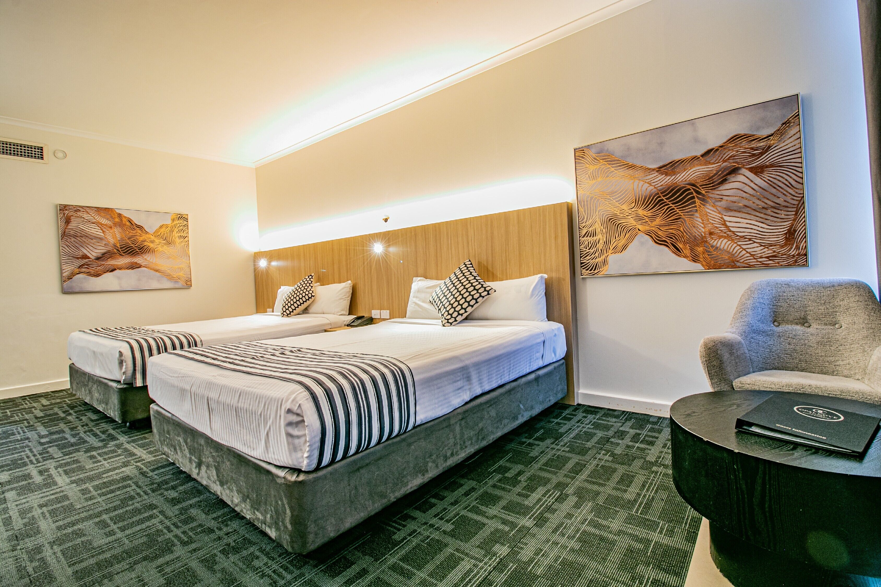 A hotel room with two beds, a wooden headboard, a green carpet, and two abstract paintings on the walls.