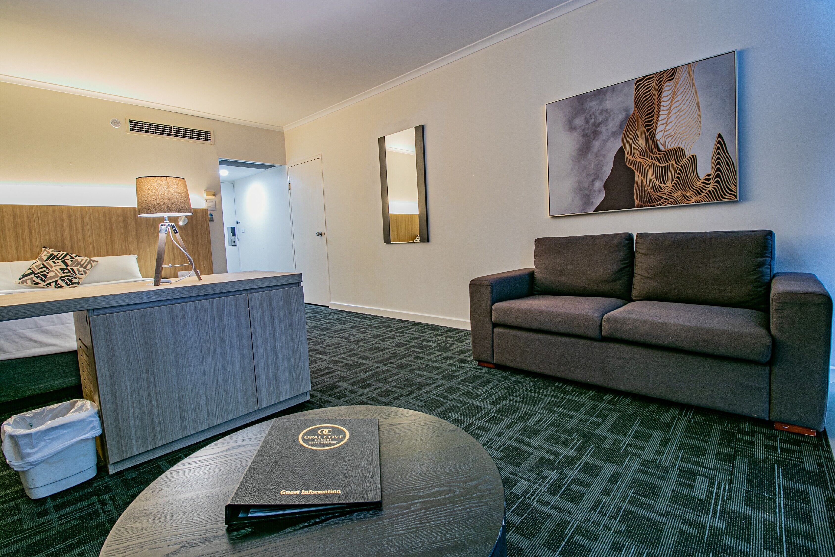 Habitació d'hotel amb un llit, una taula de treball amb una làmpada, un sofà, una taula rodona amb una carpeta i un quadre a la paret.