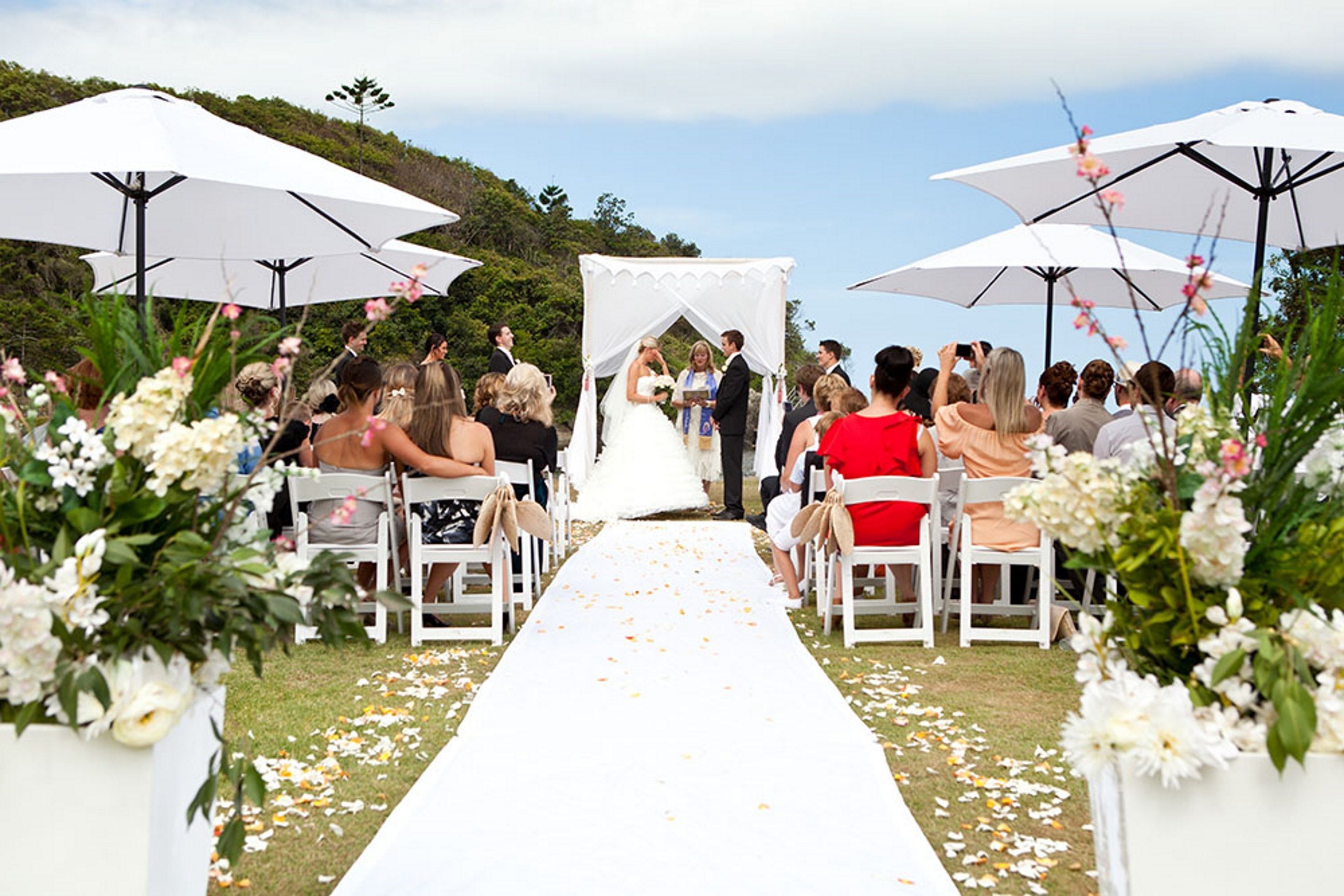A wedding ceremony with a white aisle, guests seated on white chairs, and white umbrellas providing shade.