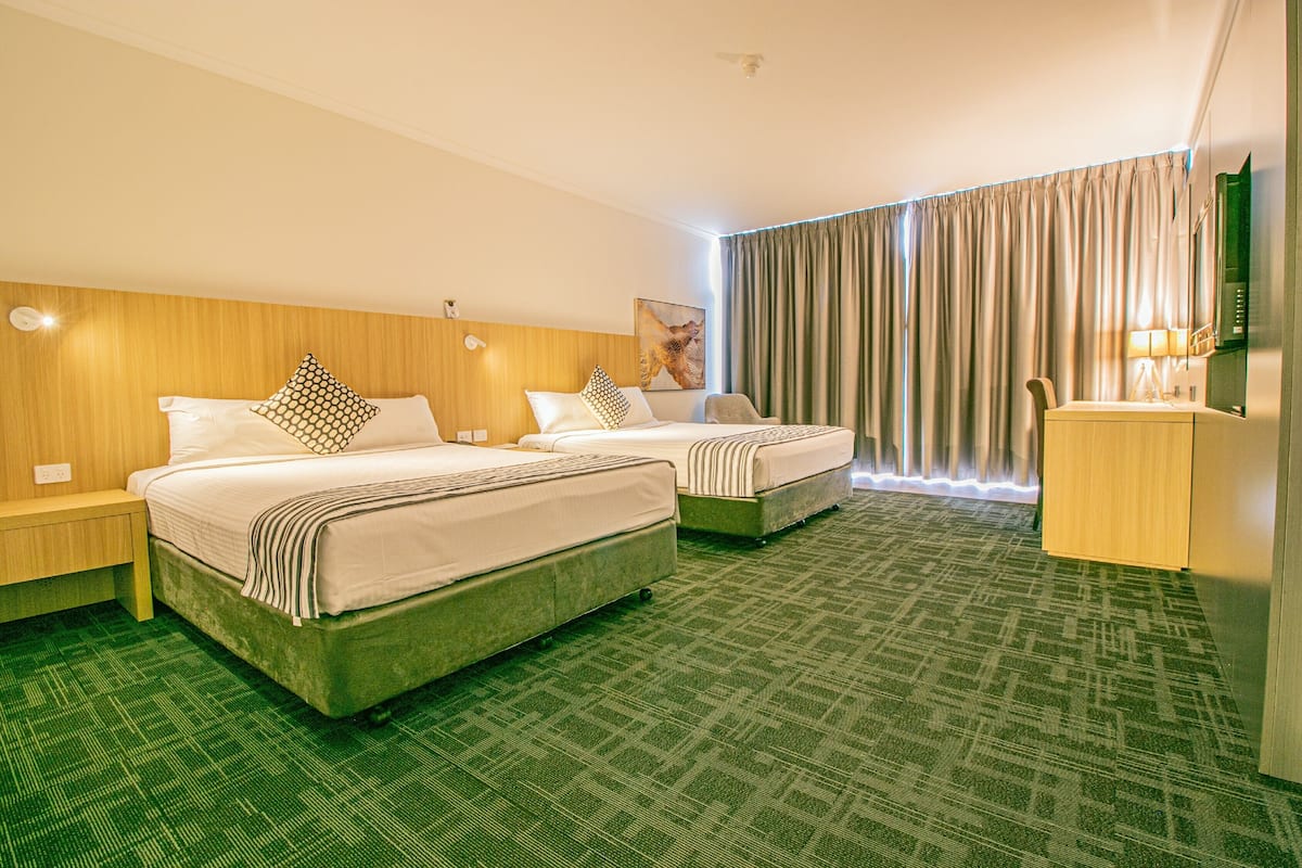 A hotel room with two beds, a wooden headboard, a green carpet, a large window with curtains, and a desk with a lamp.