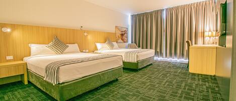 Habitación de hotel con dos camas, cabecero de madera, alfombra verde, ventana grande con cortinas y un escritorio con lámpara.