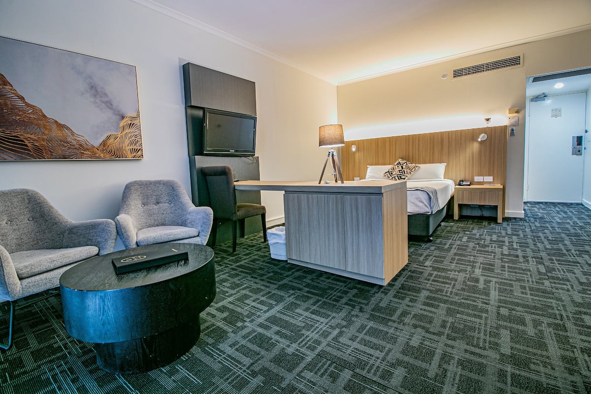 A hotel room with a bed, a desk, two armchairs, a TV, and a painting on the wall.
