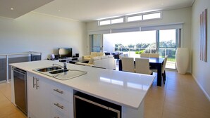 A modern kitchen with a central island, a dining area with a table and chairs, and a living space with a sofa and TV. - Opal Cove Resort (Korora)