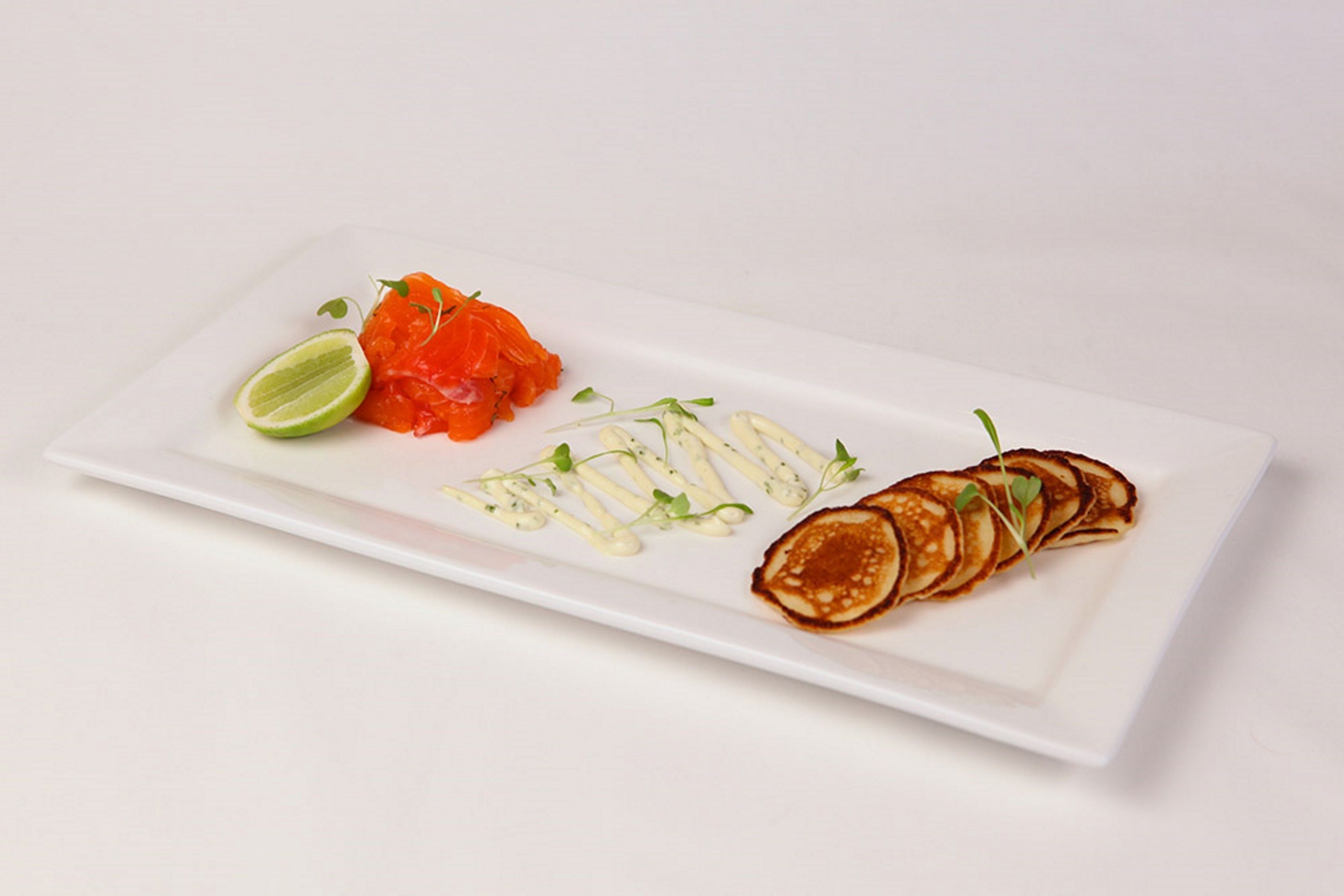 Un plato rectangular blanco con una porción de salmón, una rodaja de lima y una pila de panqueques con salsa y decoración.