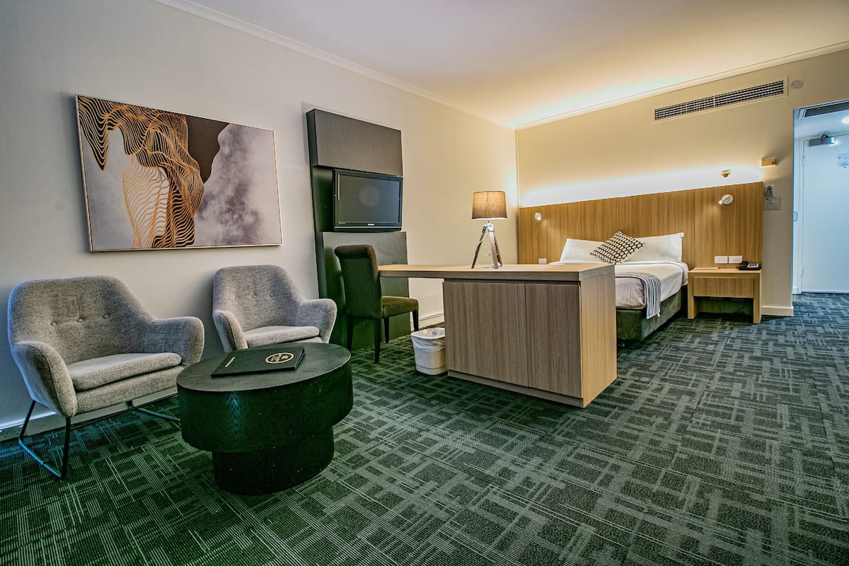 A hotel room with a bed, two armchairs, a desk, a TV, and a painting on the wall.