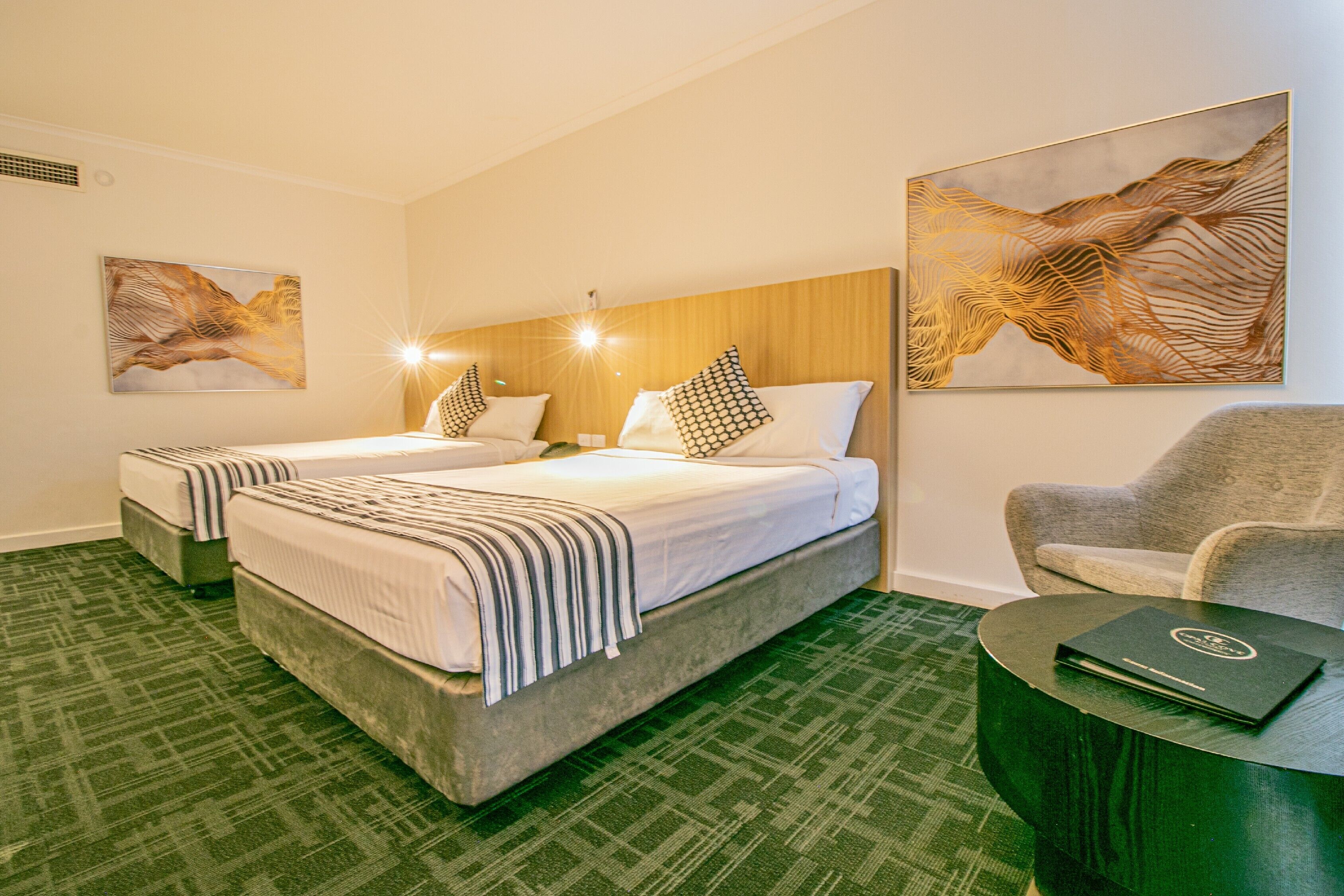 Quarto de hotel com duas camas, um tapete verde, cabeceira de madeira, uma poltrona cinza e uma mesa redonda com um laptop.