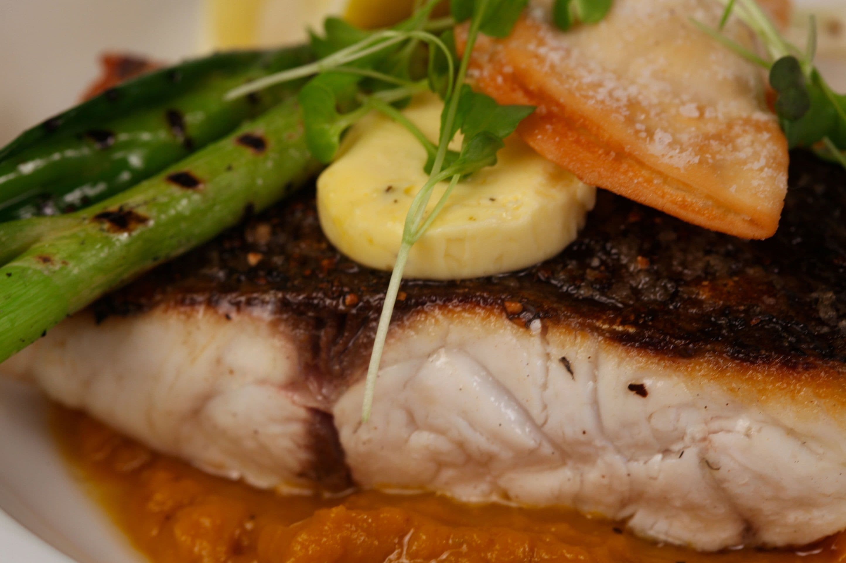 A close-up of a grilled fish fillet with a golden-brown crust, accompanied by asparagus and a slice of orange fruit, possibly a sweet potato, on a bed of sauce.