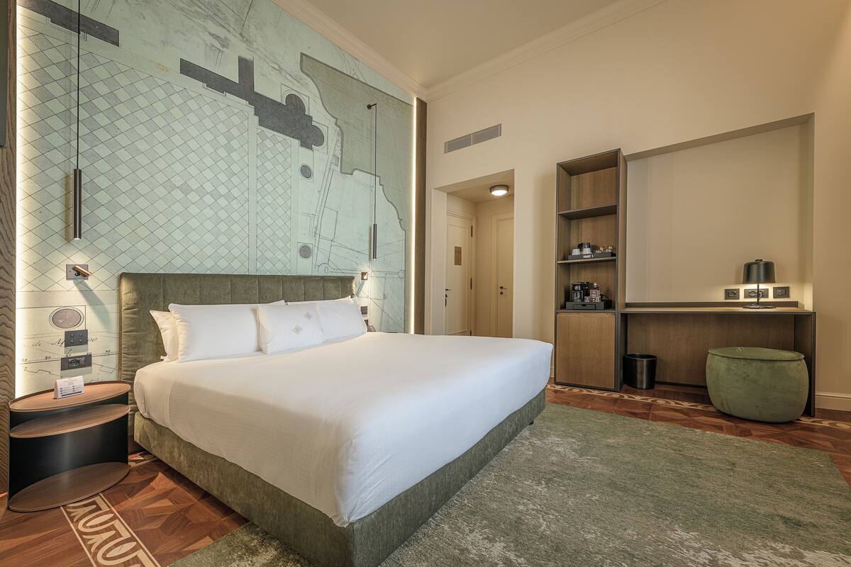 A neatly arranged hotel room with a large bed, a small side table, a green ottoman, a wooden cabinet, and a map on the wall.