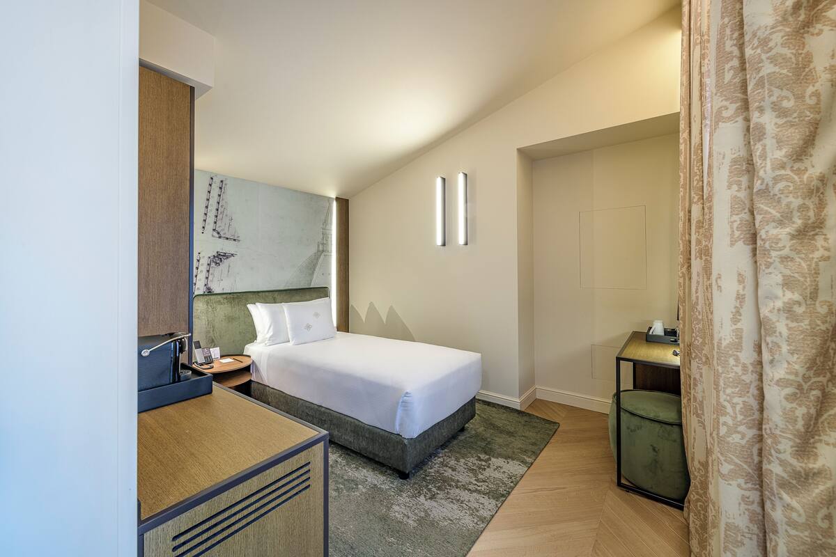 A hotel room with a bed, a desk, a chair, a nightstand, a painting on the wall, and a door.