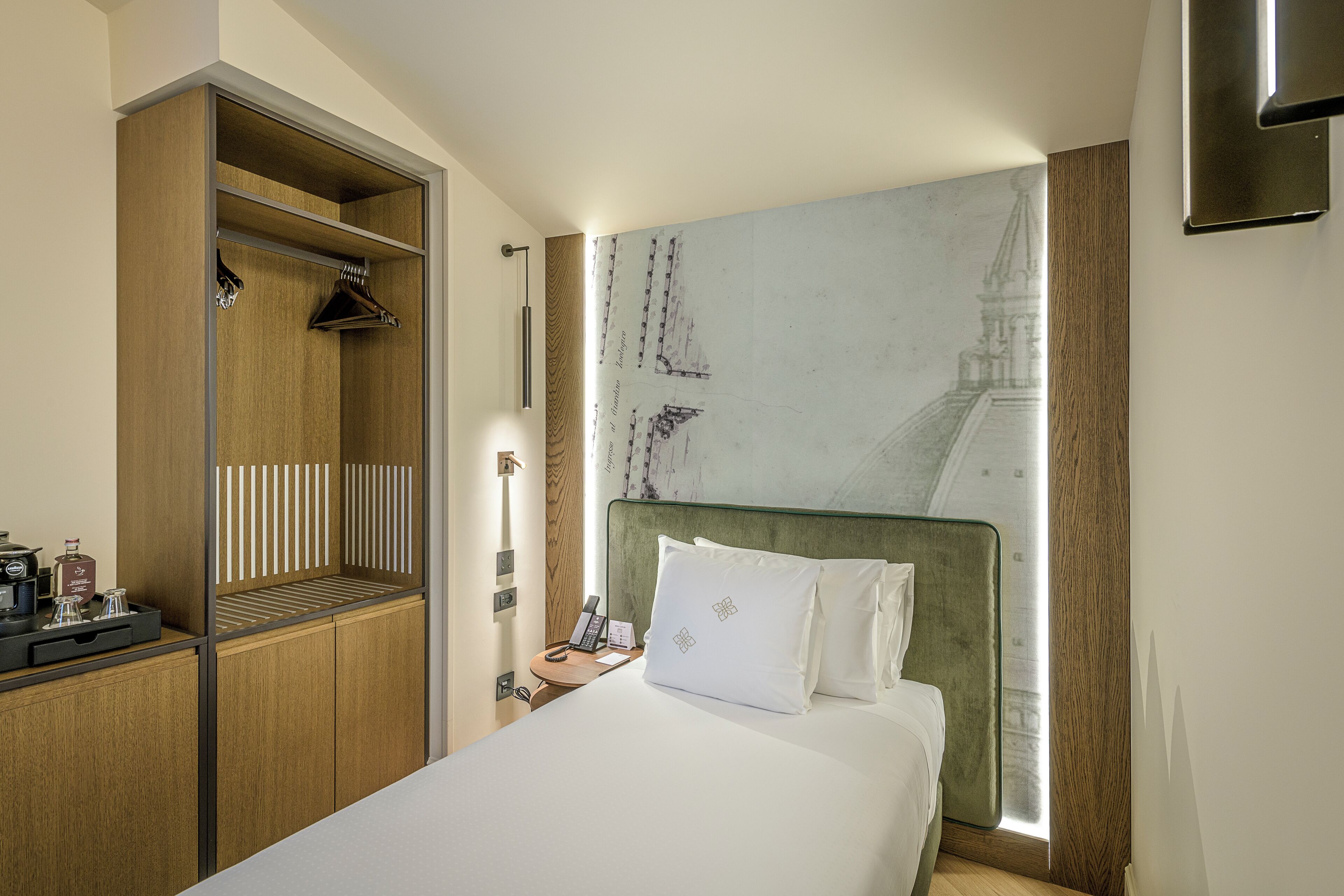 A hotel room with a bed, a wooden wardrobe, a nightstand with a lamp, and a wall-mounted artwork.