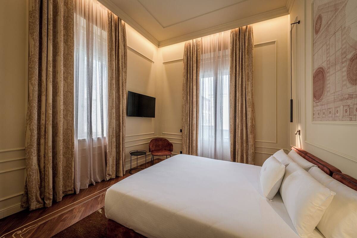 A hotel room with a large bed, curtains, a flat-screen TV, a small chair, and a wall-mounted artwork.