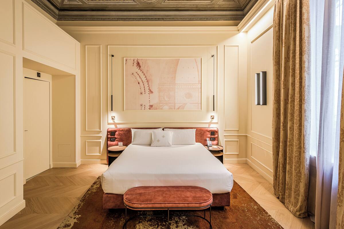 A hotel room with a large bed, a bench, a painting on the wall, and a view of the city through the window.