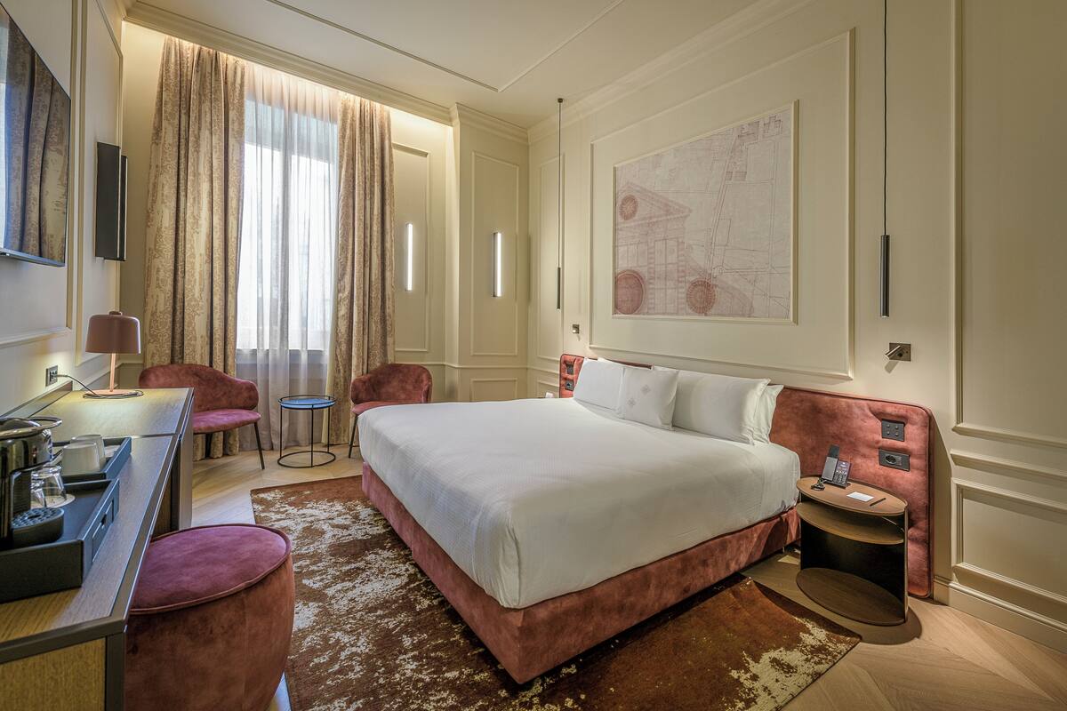 A hotel room with a large bed, a desk with a lamp, a chair, a small table, and a map on the wall.