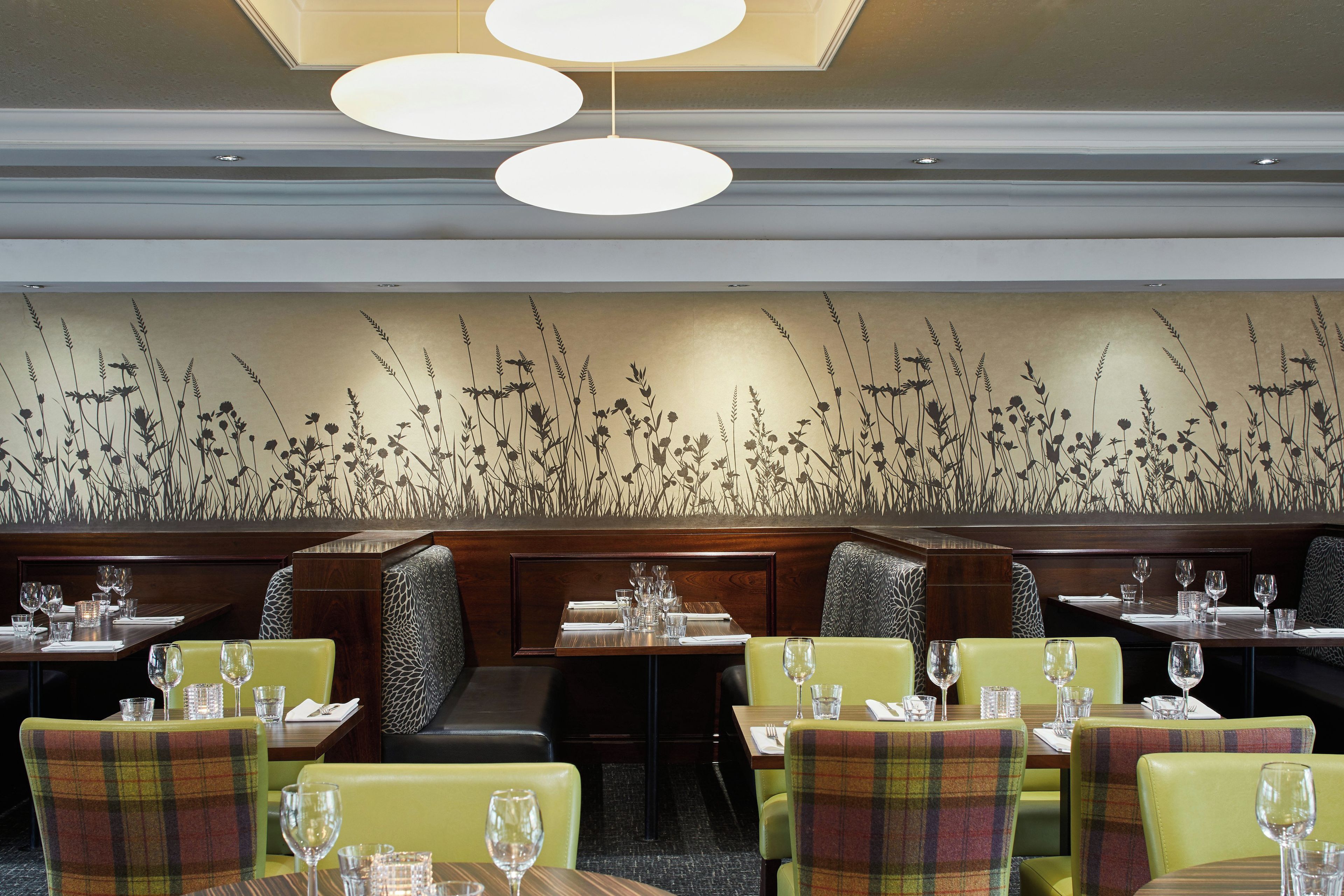 L’intérieur d’un restaurant présente des tables hautes, des chaises recouvertes d’un tissu vert et un mur décoratif orné de hautes herbes.