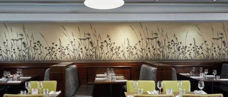 Un interior de restaurante con mesas altas, sillas tapizadas en verde y una pared decorativa con pastos altos.