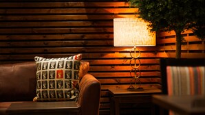 Een knus vertrek met een wand van houten latten, een met kussens beklede bank, een gestreept kussen, een tafel met een lamp en een potplant.
