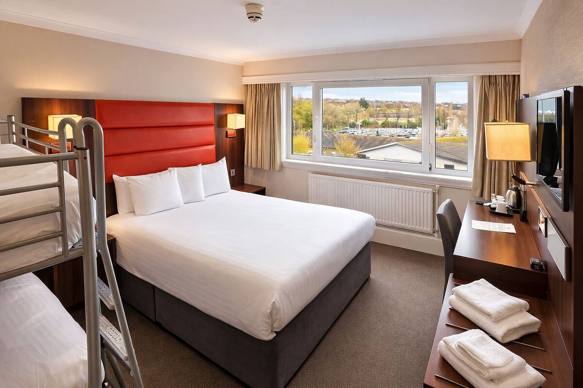 A hotel room with a bunk bed, a desk with a chair, a TV, and a window with a view of a parking lot and buildings.