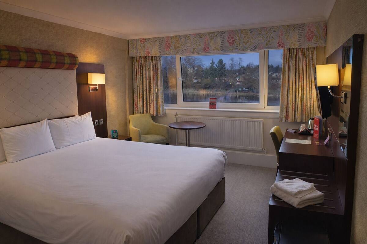 A hotel room with a large bed, a desk, a chair, a small table, and a window with curtains.