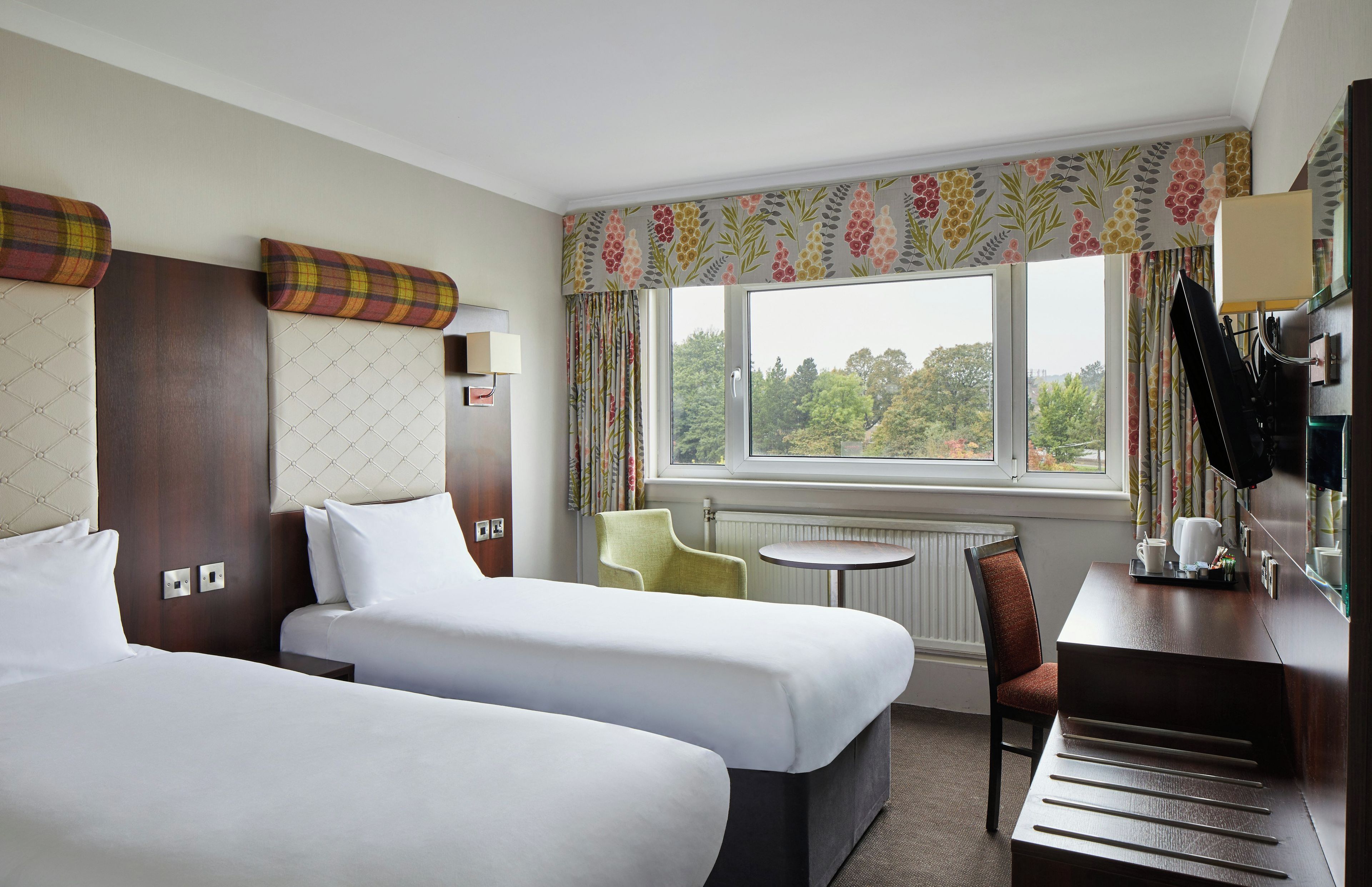 A hotel room with two beds, a small table, a chair, and a window with a view of trees.