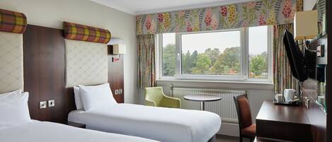 A hotel room with two beds, a small table, a chair, and a window with a view of trees.