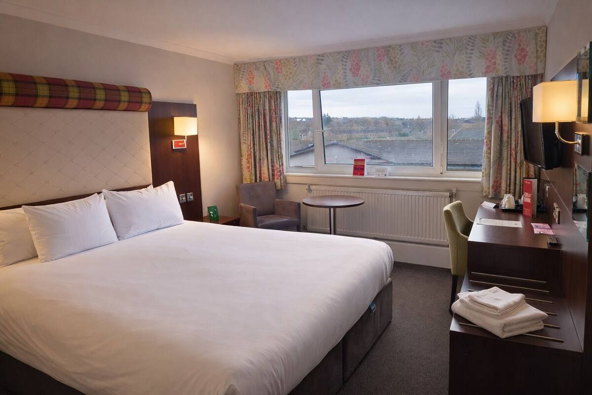 A hotel room with a large bed, a desk, a chair, a small table, and a view of the city through the window.