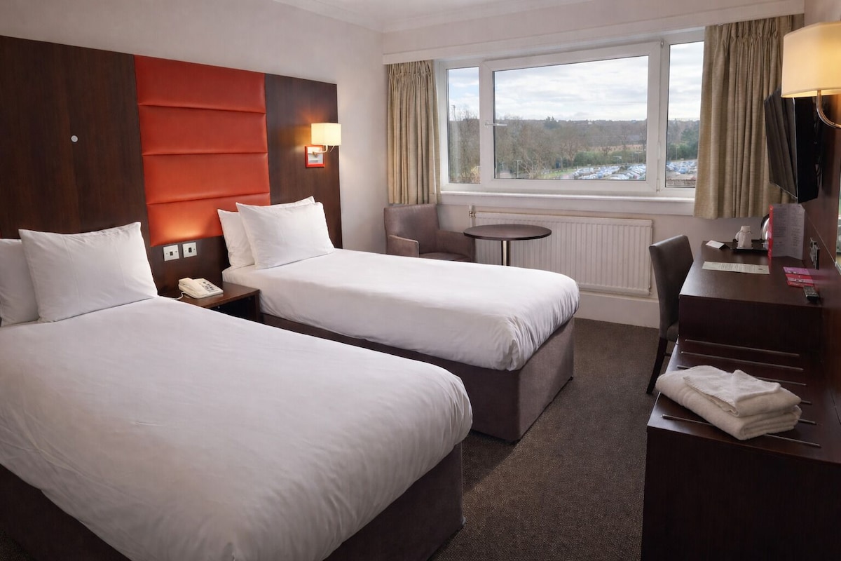 A hotel room with two beds, a desk, a chair, a small table, a lamp, and a window with a view.