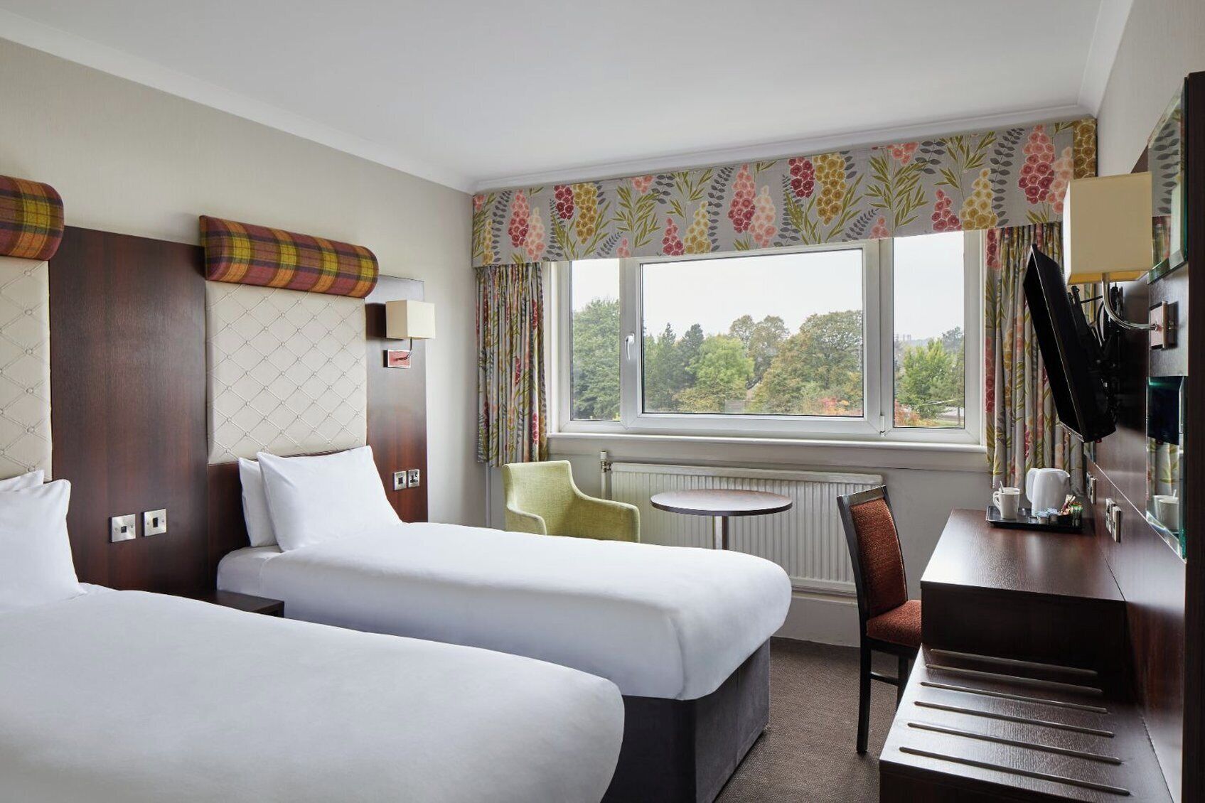 Een hotelkamer met twee bedden, een tafeltje, een stoel, een bureau met spiegel en een raam met uitzicht op bomen.