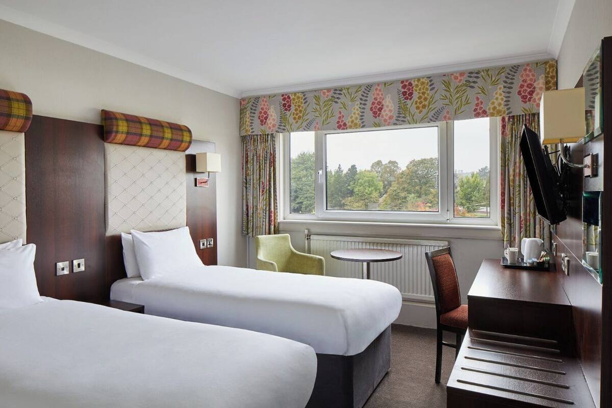 A hotel room with two beds, a small table, a chair, a desk with a mirror, and a window with a view of trees.