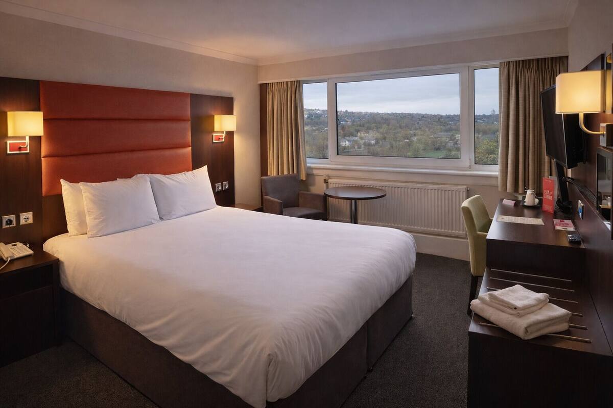 A hotel room with a large bed, a desk with a chair, a small table, and a view of the outside.
