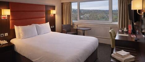 A hotel room with a large bed, a desk with a chair, a small table, and a view of the outside.