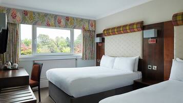 A hotel room with two beds, a desk with a chair, a window with curtains, and a view of trees.