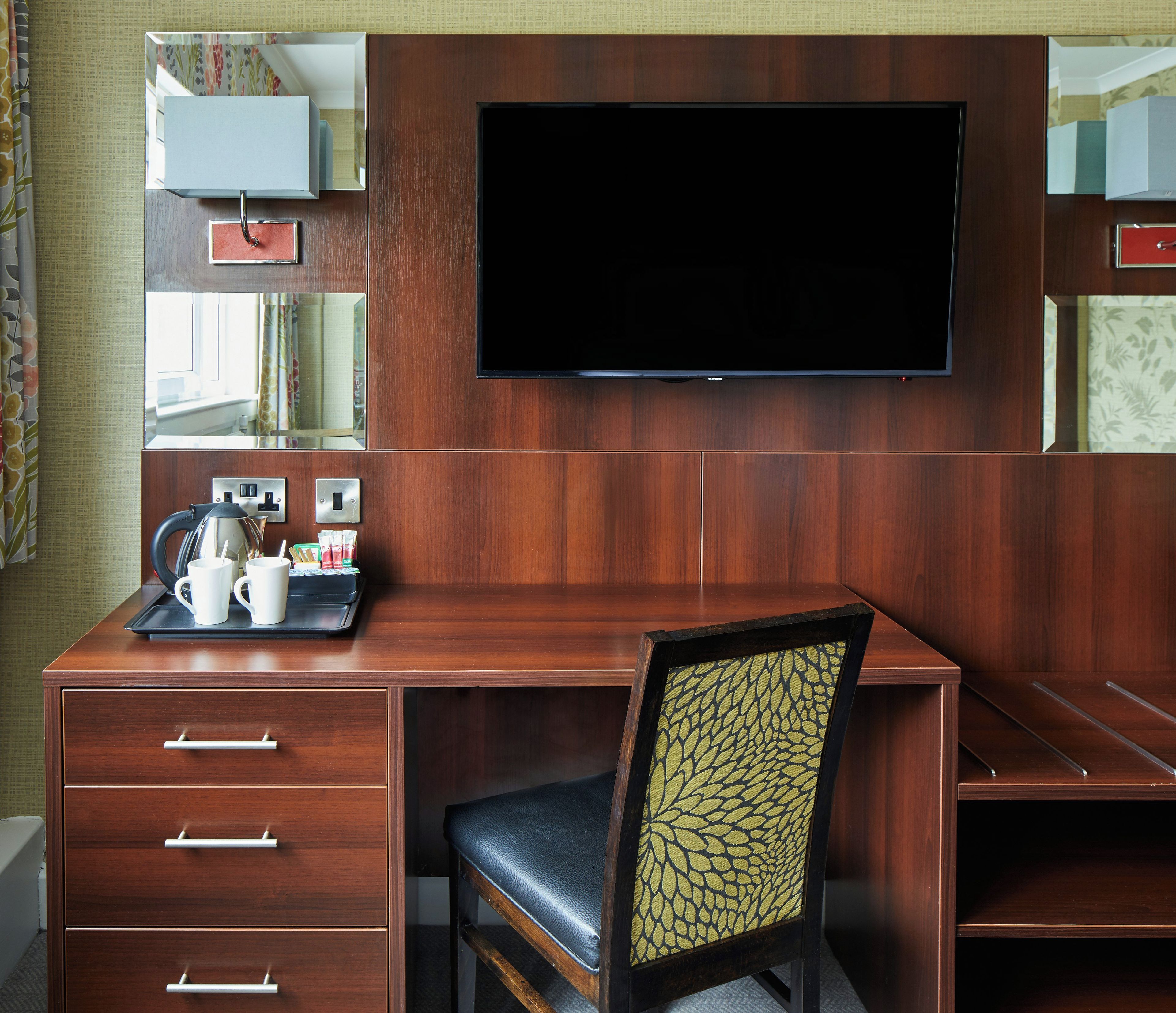A hotel room with a wooden desk, a flat-screen TV mounted on the wall, a chair with a patterned cushion, and a tray with a coffee pot and cups.