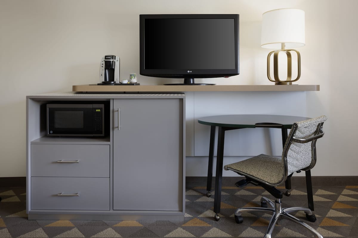 Una habitación de hotel con escritorio, monitor de computadora, lámpara, microondas y una silla.