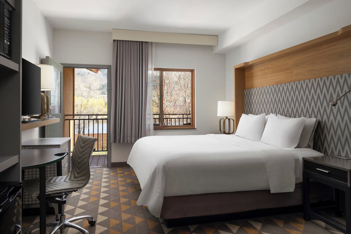 Una habitación de hotel con cama, escritorio, silla y vistas a árboles por la ventana.
