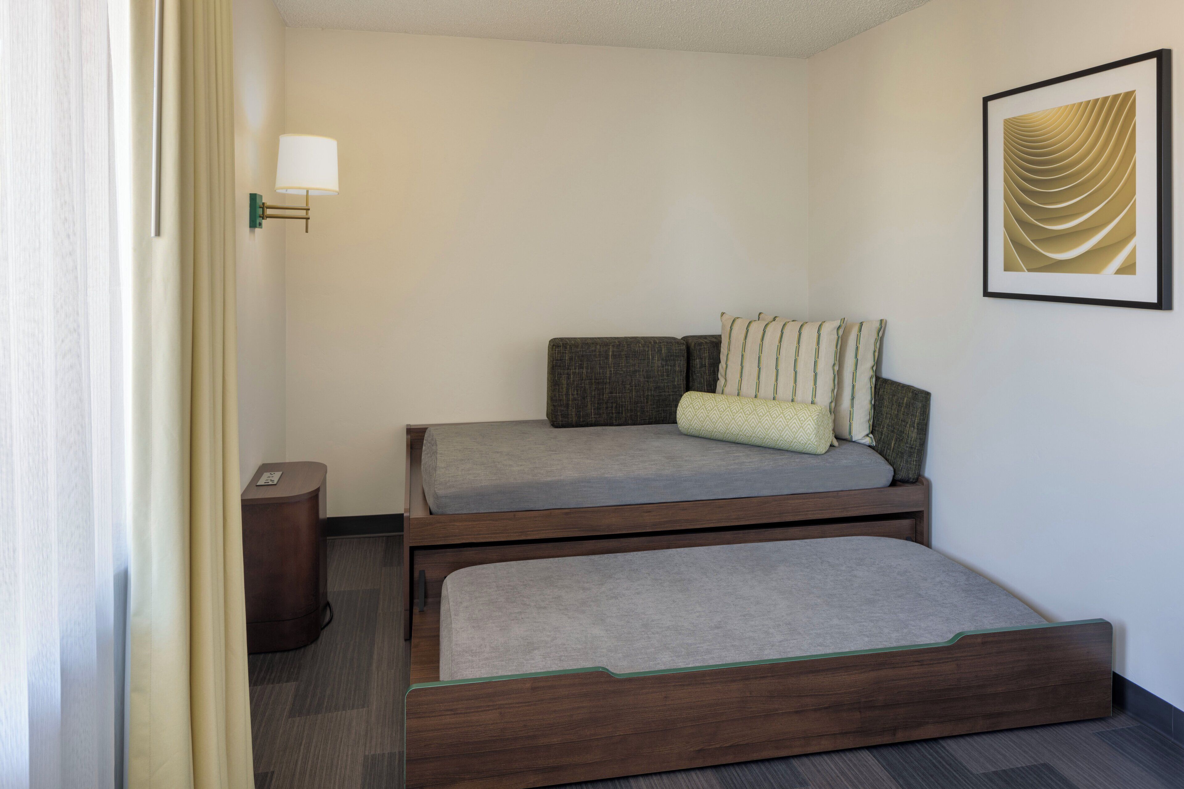 Ein kleines Hotelzimmer mit Bett, Nachttisch, Lampe und einem gerahmten Bild an der Wand.