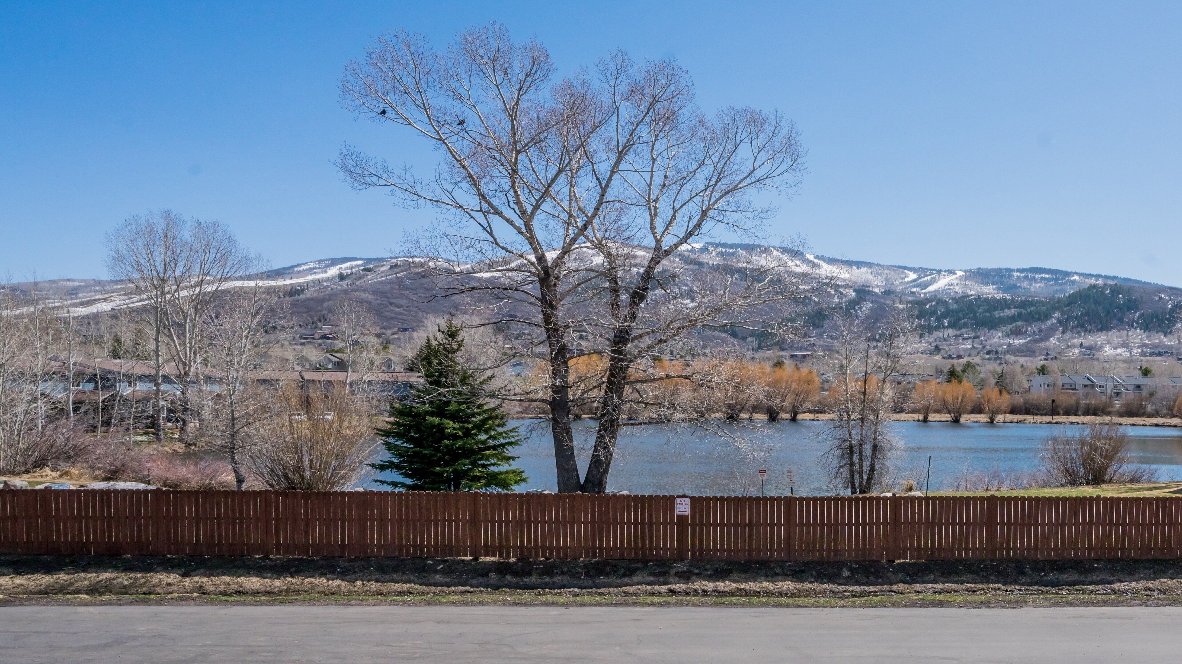 A view of a lake with a mountain in the background, a wooden fence in the foreground, and a clear blue sky.