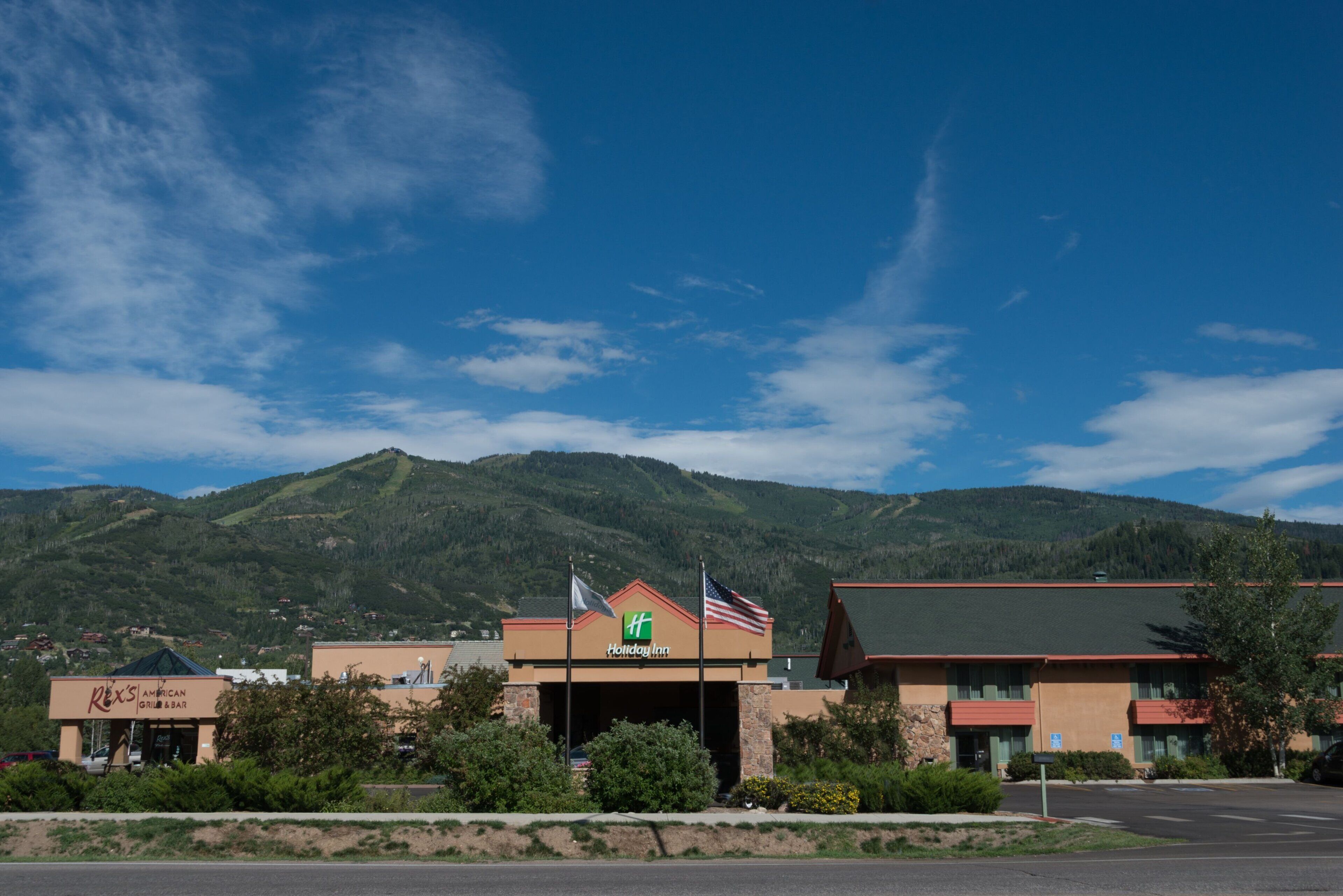 A Holiday Inn with a mountain backdrop, a Rex's American Grill & Bar, and a parking lot.