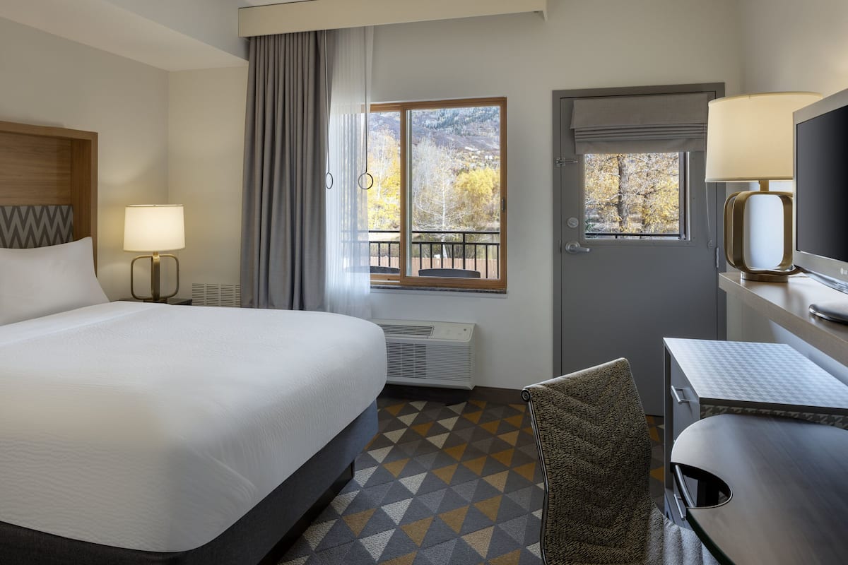 Habitación de hotel con una cama grande, un escritorio con silla, una ventana con vista a árboles y una puerta.