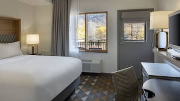 A hotel room with a large bed, a desk with a chair, a window with a view of trees, and a door.