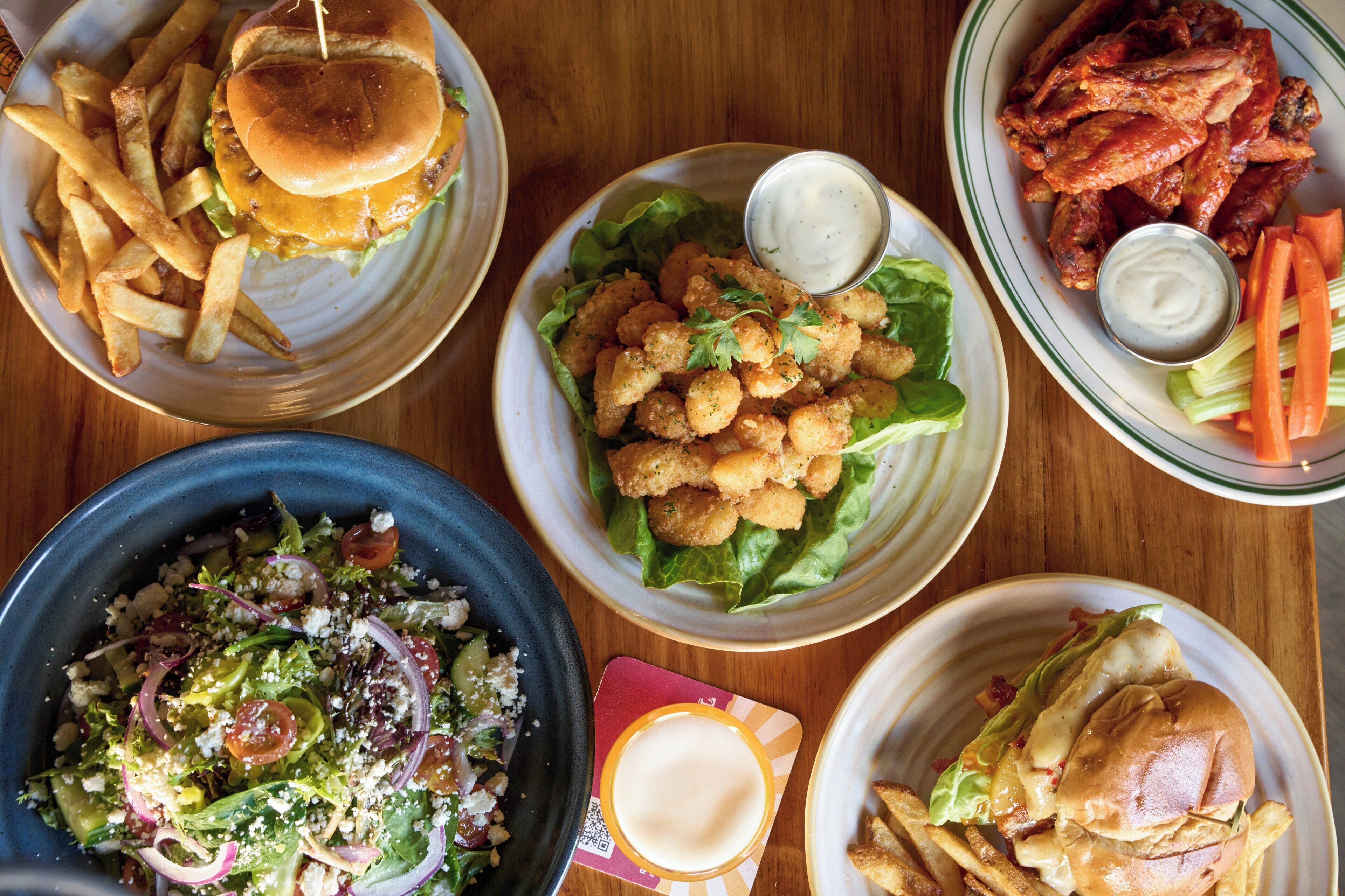 Variedad de platos, como hamburguesas, papas fritas, ensalada y alitas, sugiriendo un ambiente de restaurante informal.