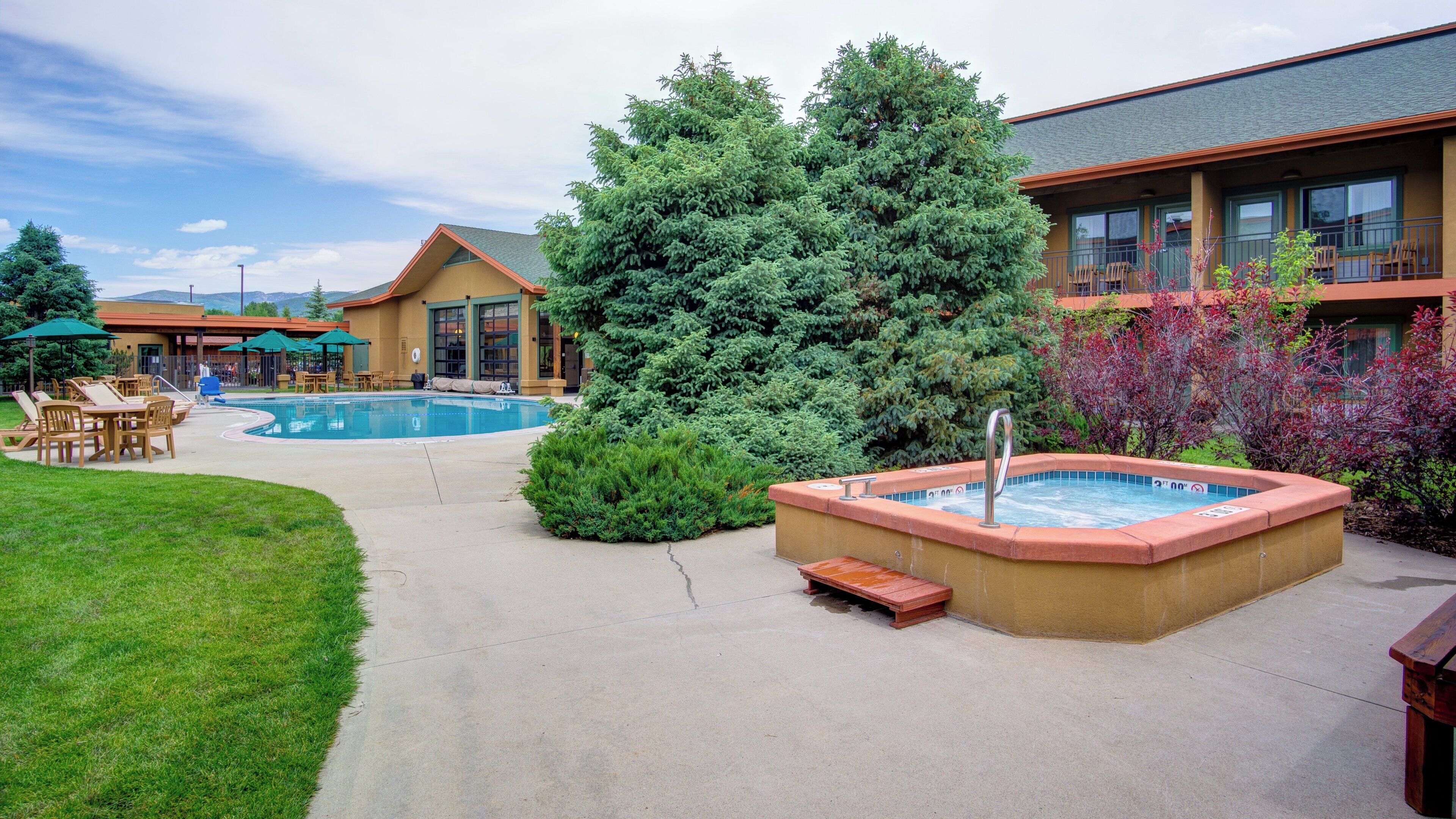 A hotel courtyard with a swimming pool, lounge chairs, a hot tub, and a large evergreen tree.
