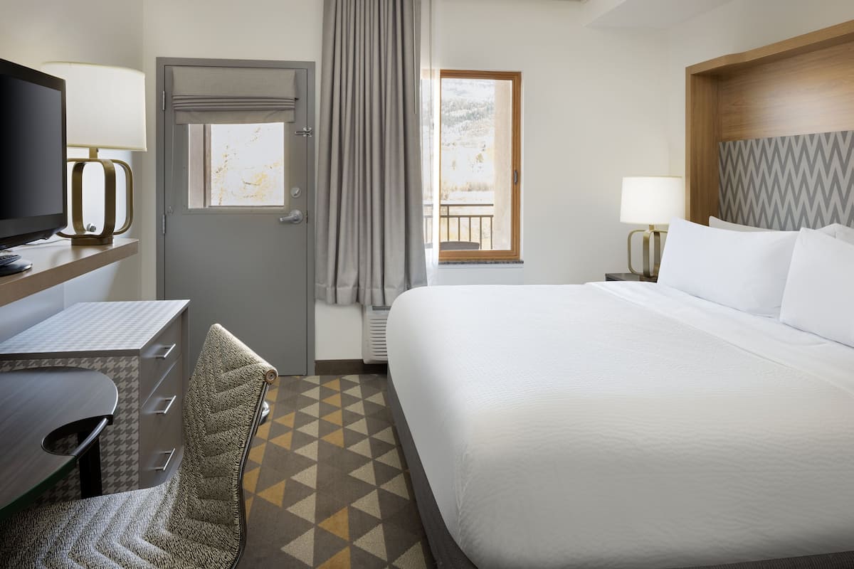 Habitación de hotel con cama, escritorio con computadora, silla, televisor y ventana con vista a un paisaje nevado.